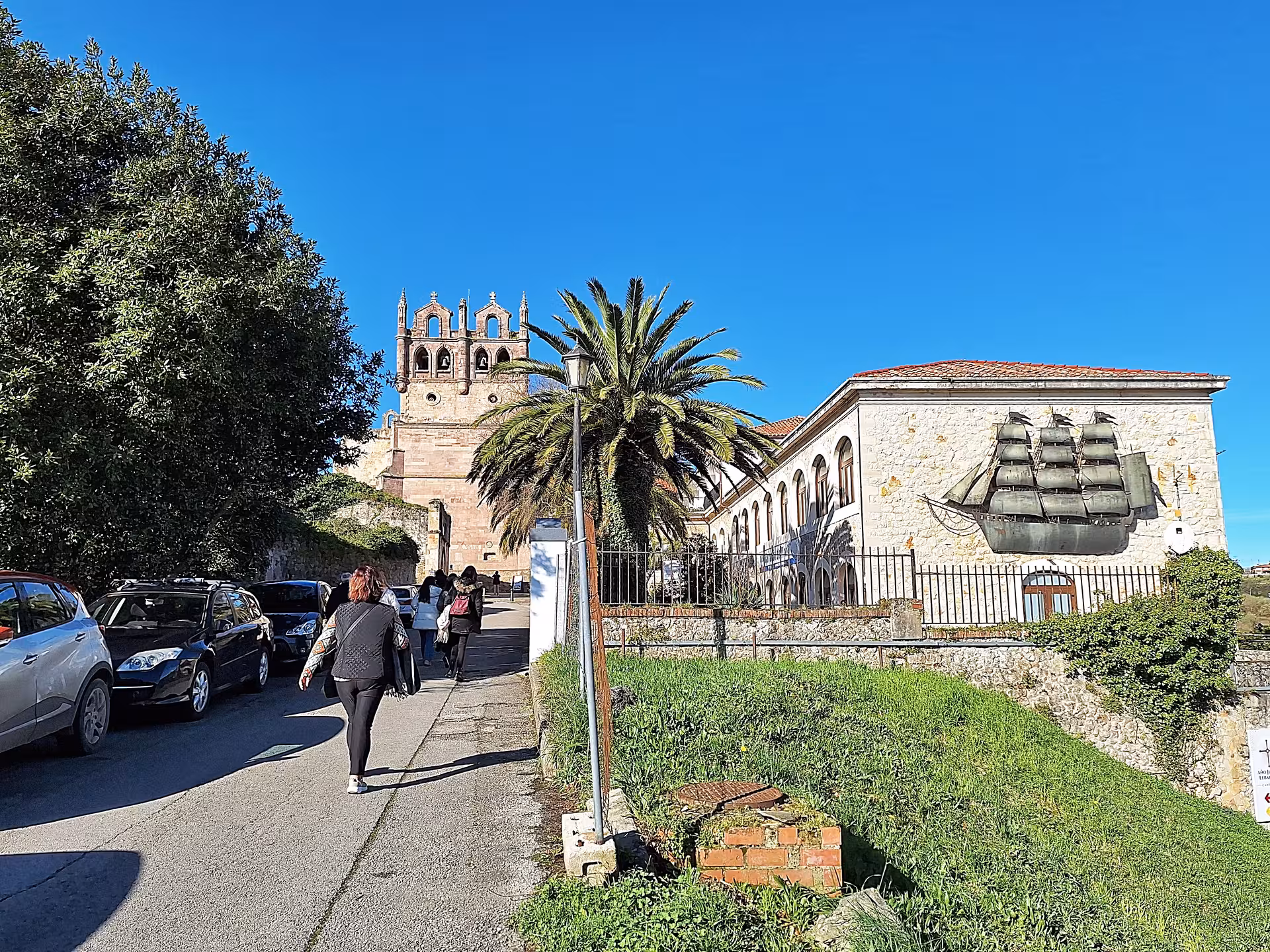 Tourists walk along a scenic path with a bell tower and mural in San Vicente de la Barquera.
