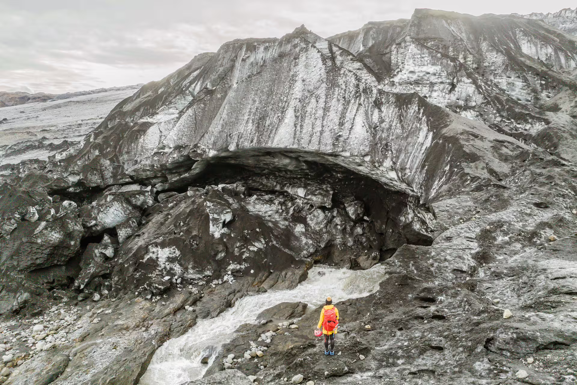 A solo hiker in a yellow jacket stands before a massive glacial ice cave arch on the Sandfellsjokull Glacier Iceland day expedition tour.