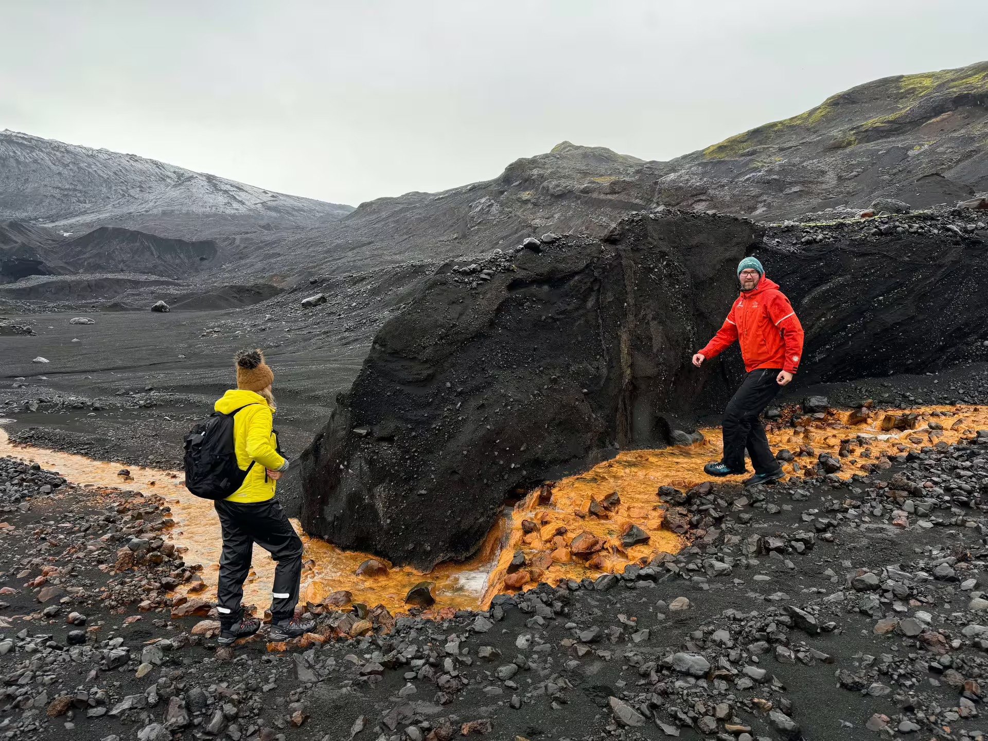 Two hikers in colorful gear explore black volcanic terrain with orange mineral streams on a Sandfellsjokull Glacier guided day trip Iceland.