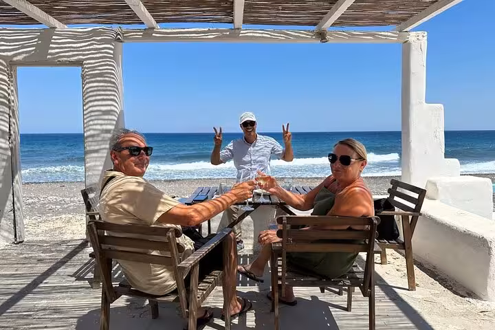 Cruise ship shore excursion in Santorini with seaside taverna stop, guests toasting by Perissa beach