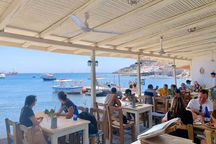 Seaside taverna stop on Santorini cruise shore tour, with diners and boats in a calm bay near the port