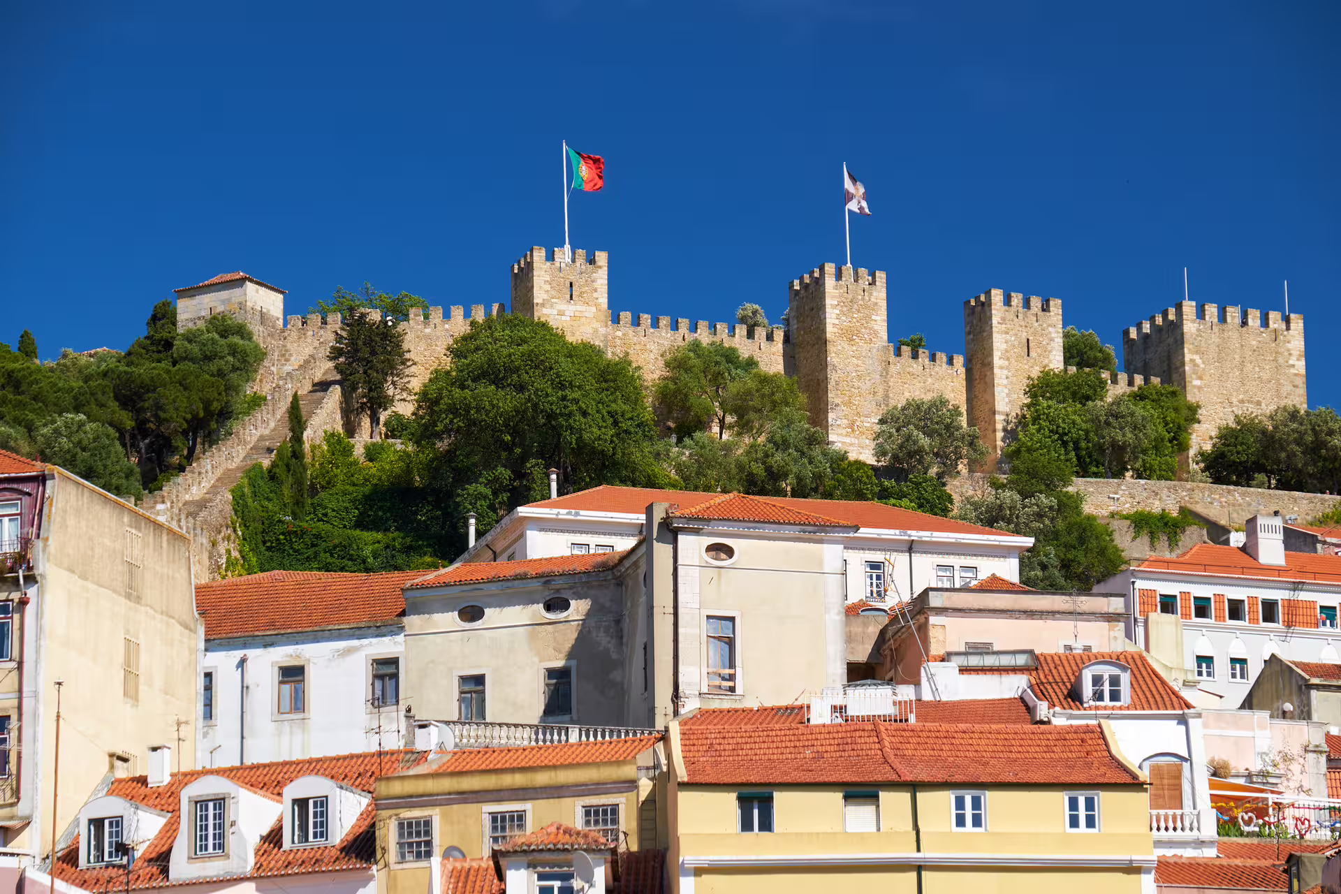 View of São Jorge Castle in Lisbon, Portugal, with red-roofed buildings below and clear blue sky, featured in shore excursion.