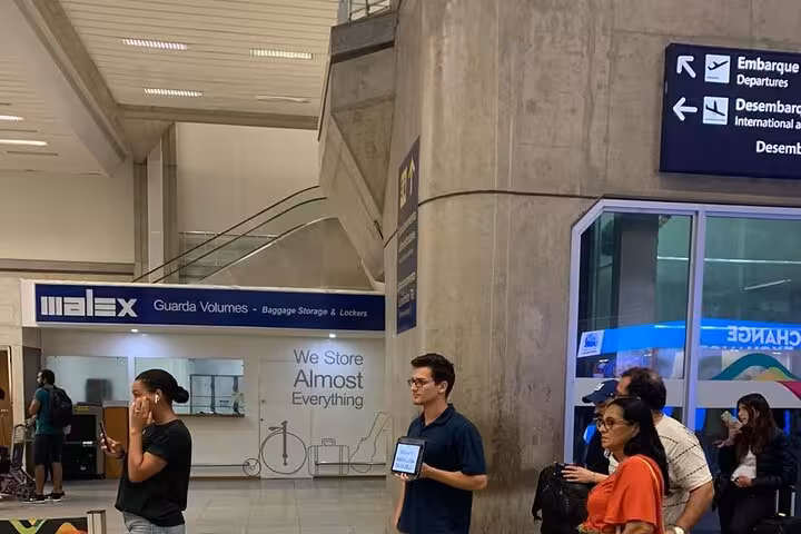 Man with a name sign waits at São Paulo airport for shuttle to Paraty, near baggage storage area.