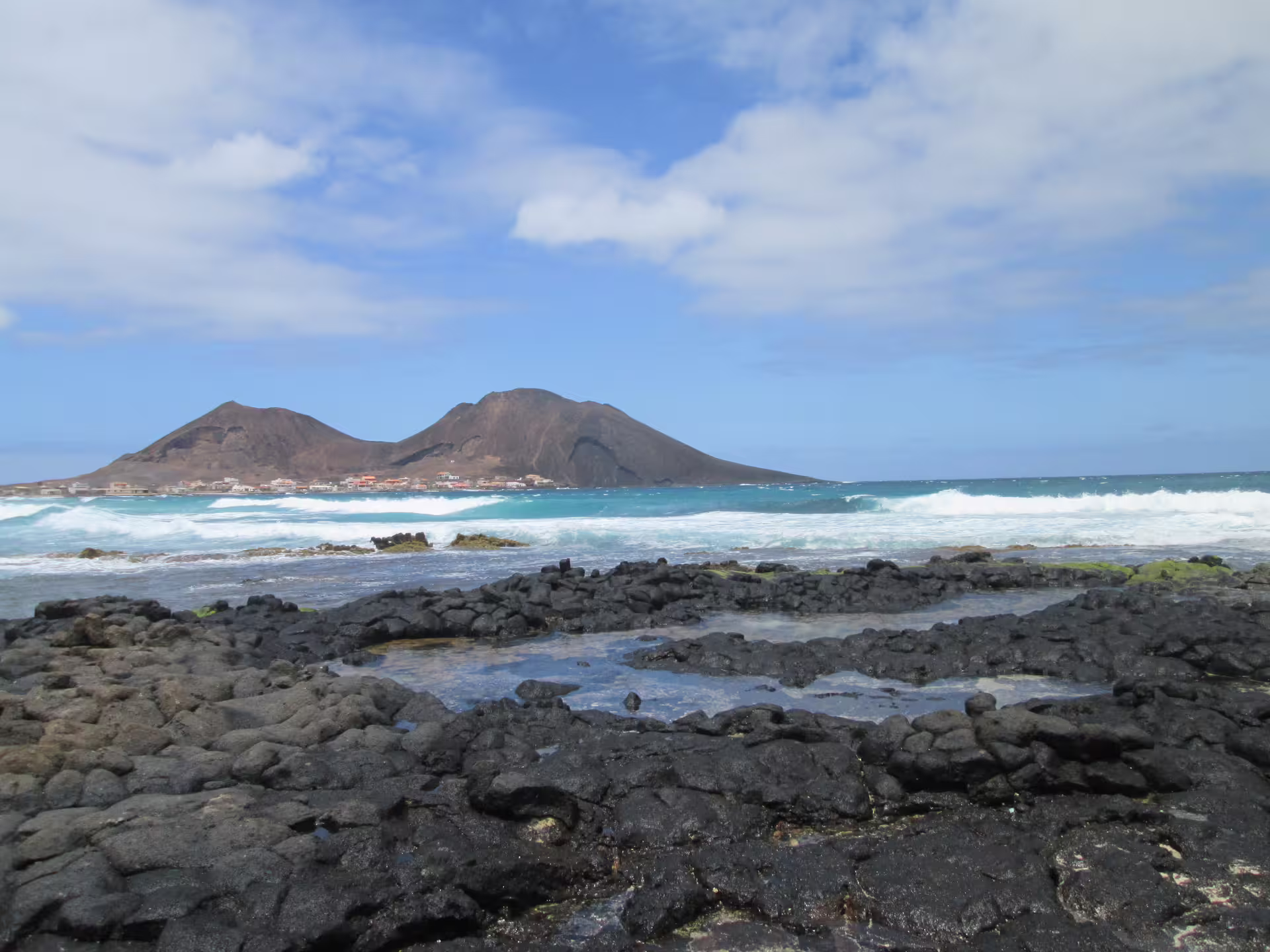 Scenic view of São Vicente's rocky coastline with turquoise waves and Monte Verde in the background under a blue sky.