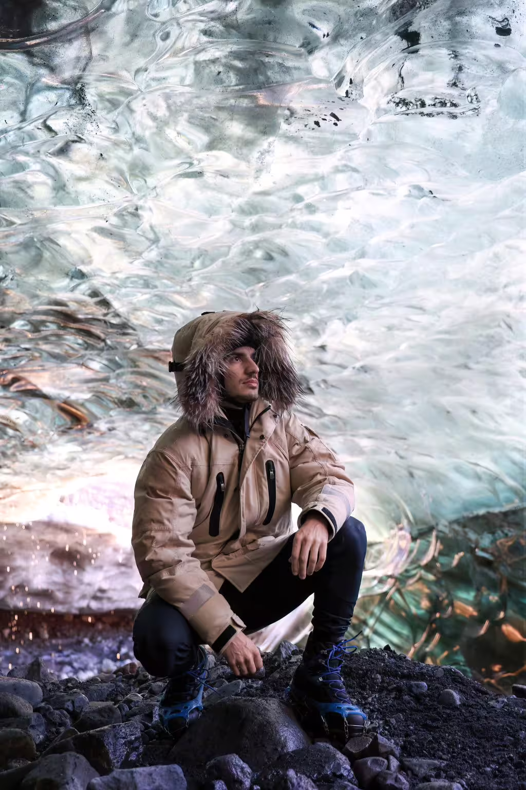 Man in hooded winter jacket sits on volcanic rocks inside a glowing sapphire ice cave on an early bird Iceland tour