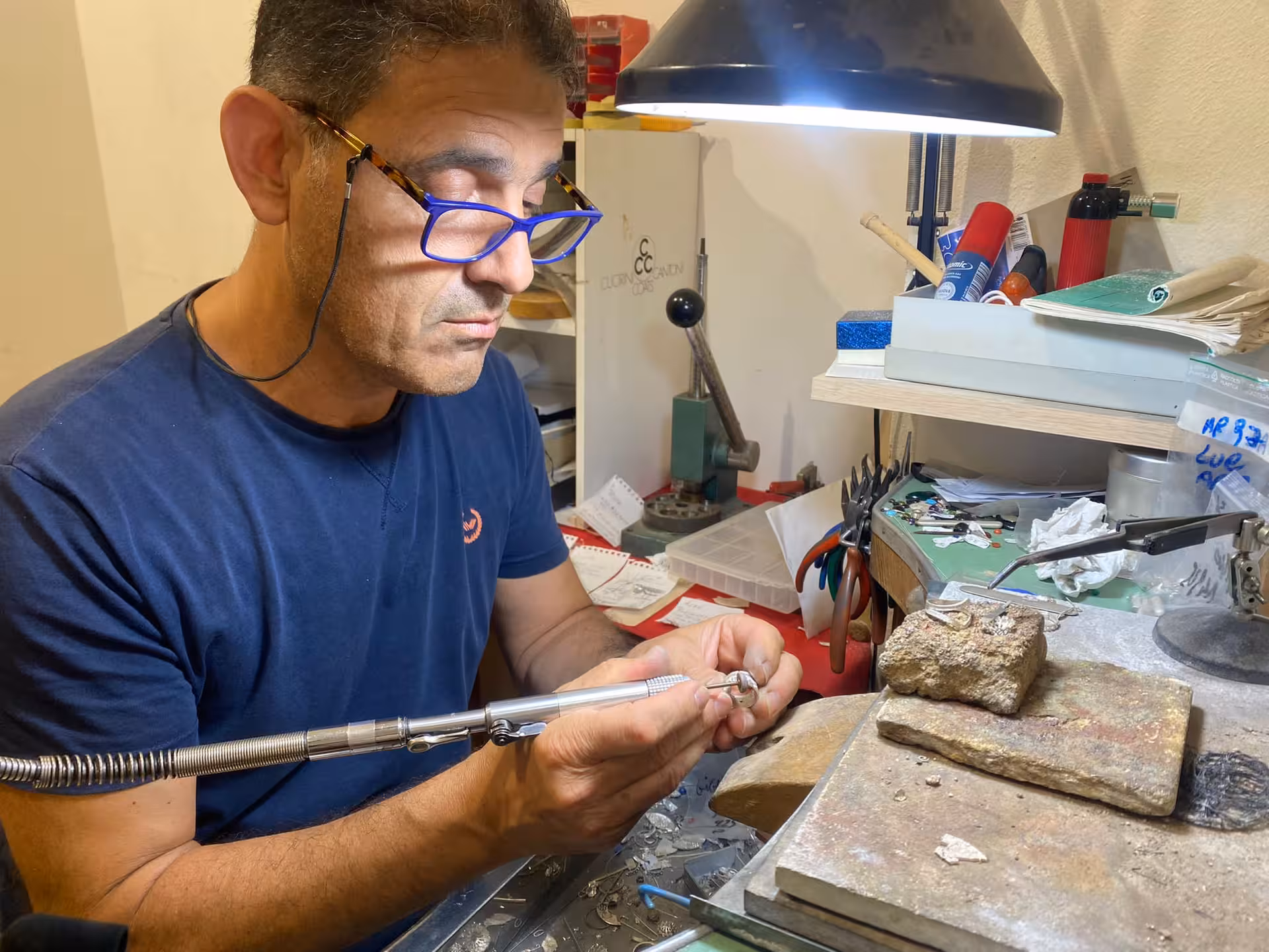 Goldsmith meticulously crafting jewelry under a lamp in a Sassari filigree workshop, surrounded by tools and materials.