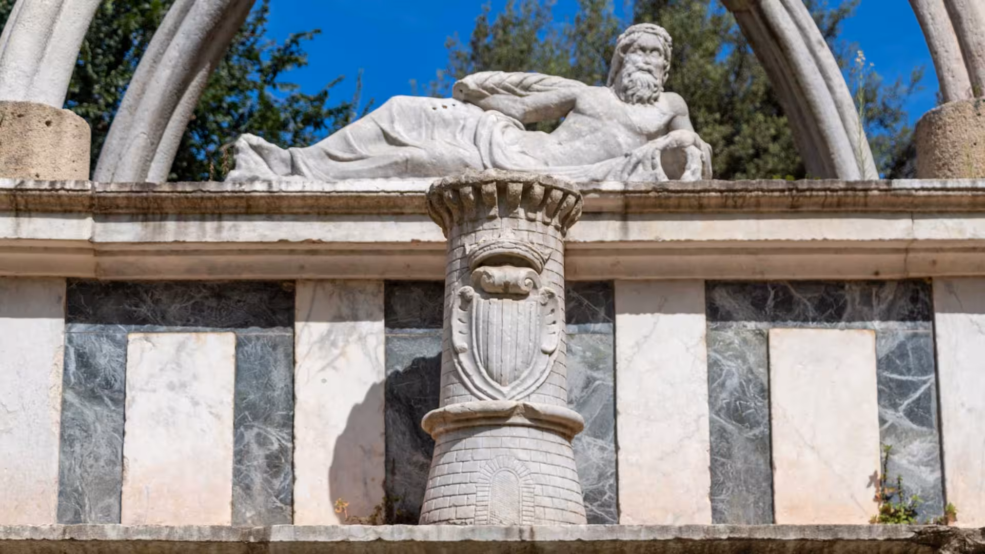 Statue of a reclining figure on a historic fountain in Sassari's city center, surrounded by lush greenery.