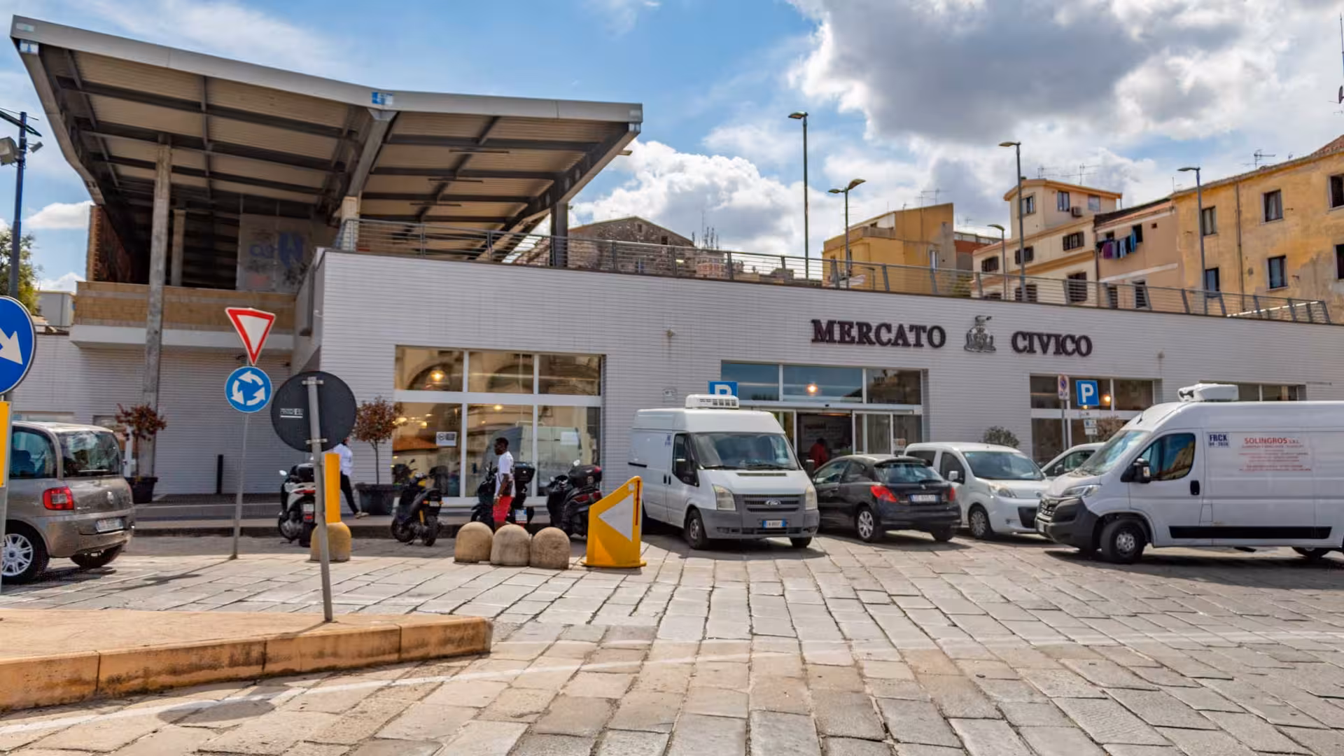 Entrance of Mercato Civico in Sassari, bustling with visitors and vehicles, a hub of local commerce and culture.