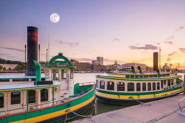 Two colorful steamboats docked on Savannah riverfront at sunset with full moon and city skyline