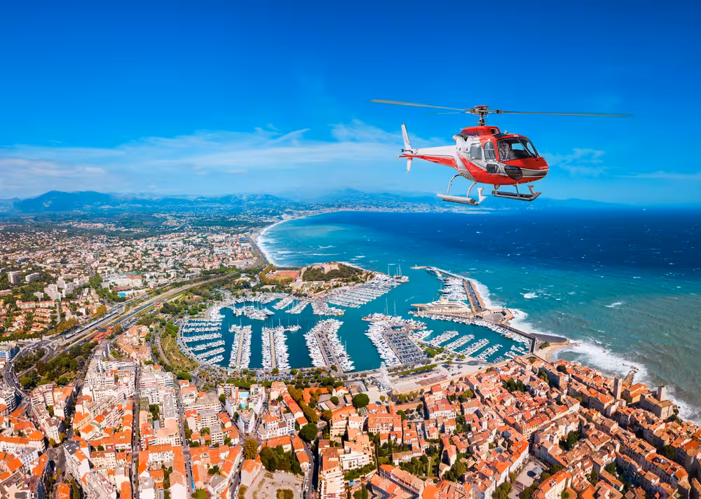 Red helicopter flying over Cannes marina and coastline with turquoise Mediterranean Sea below