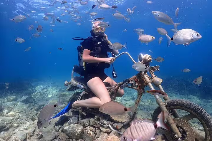 Scuba diver poses on a submerged motorcycle surrounded by fish during an exciting Kusadasi dive tour.