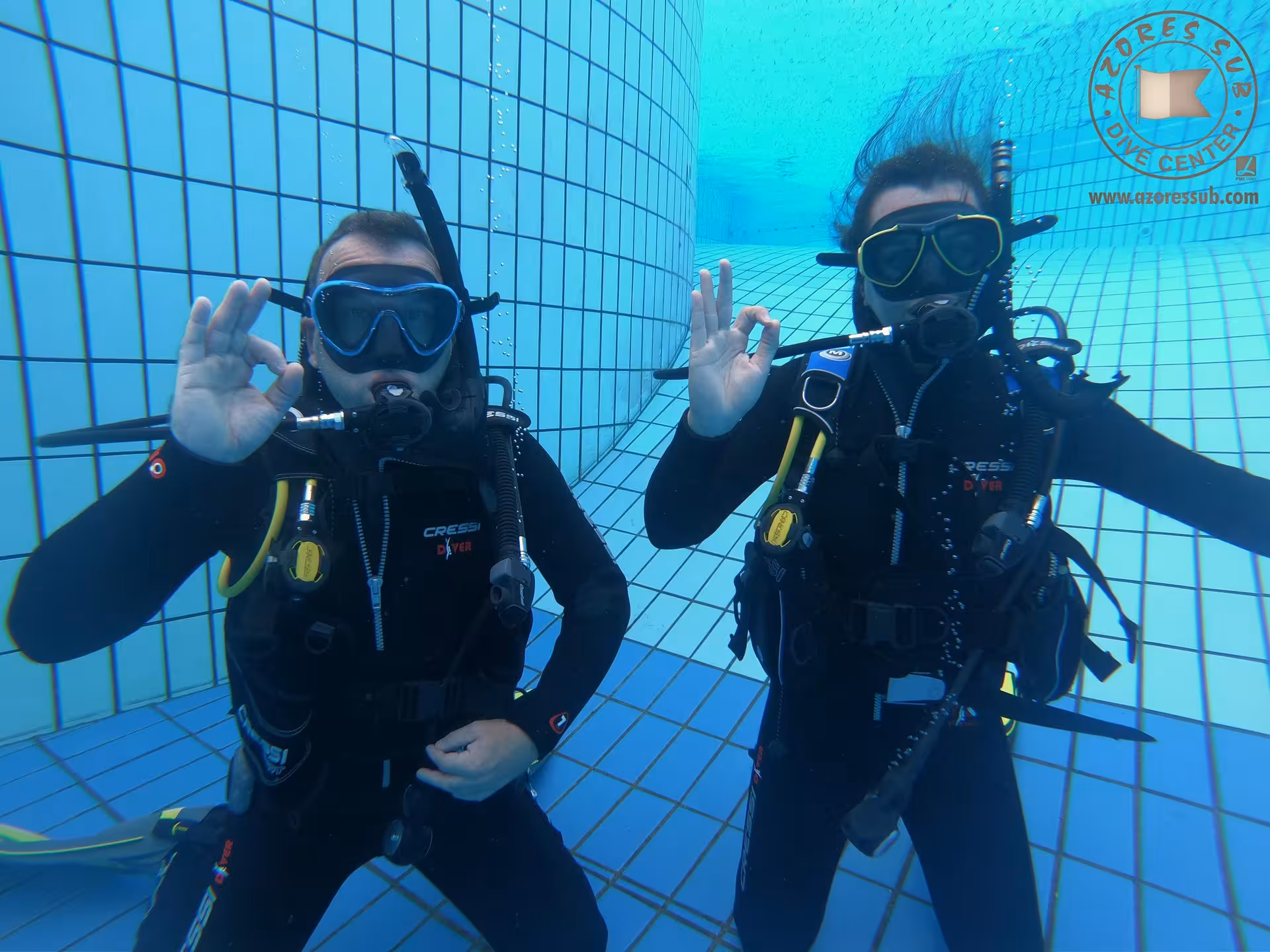 Two scuba divers give OK sign underwater in tiled pool during baptism-style swimming pool dive experience