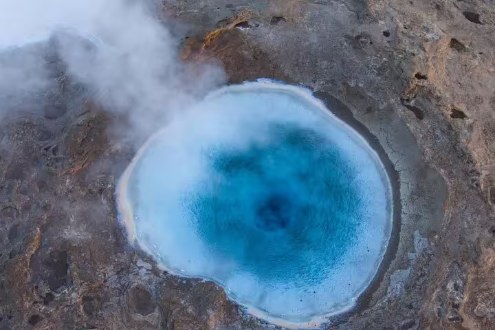 Aerial view of a vivid blue geothermal hot spring pool with steam rising from rocky landscape