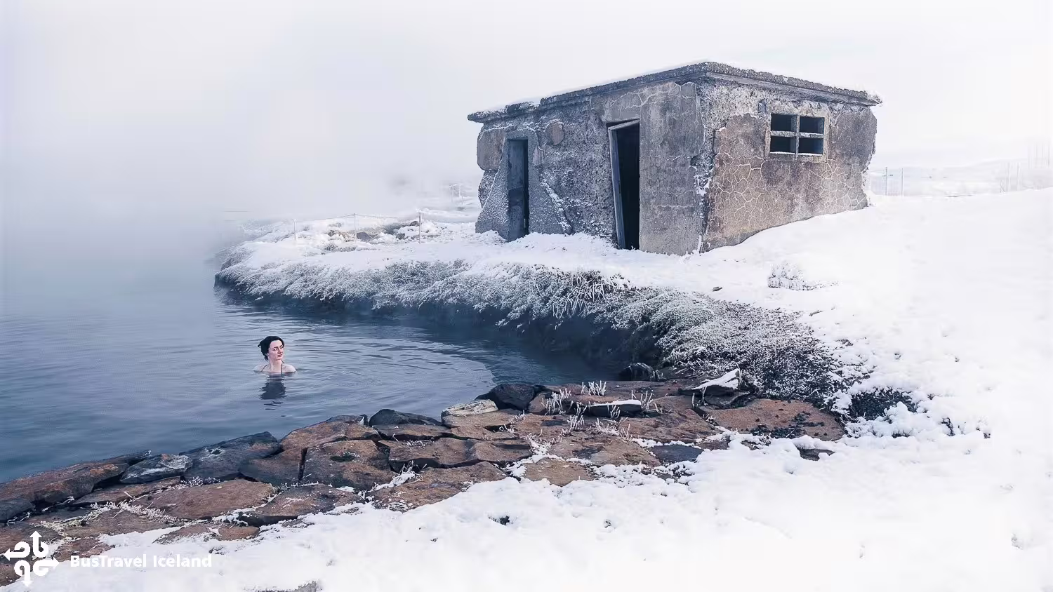 Woman bathing in Secret Lagoon geothermal hot spring surrounded by snow and old stone changing hut, Iceland