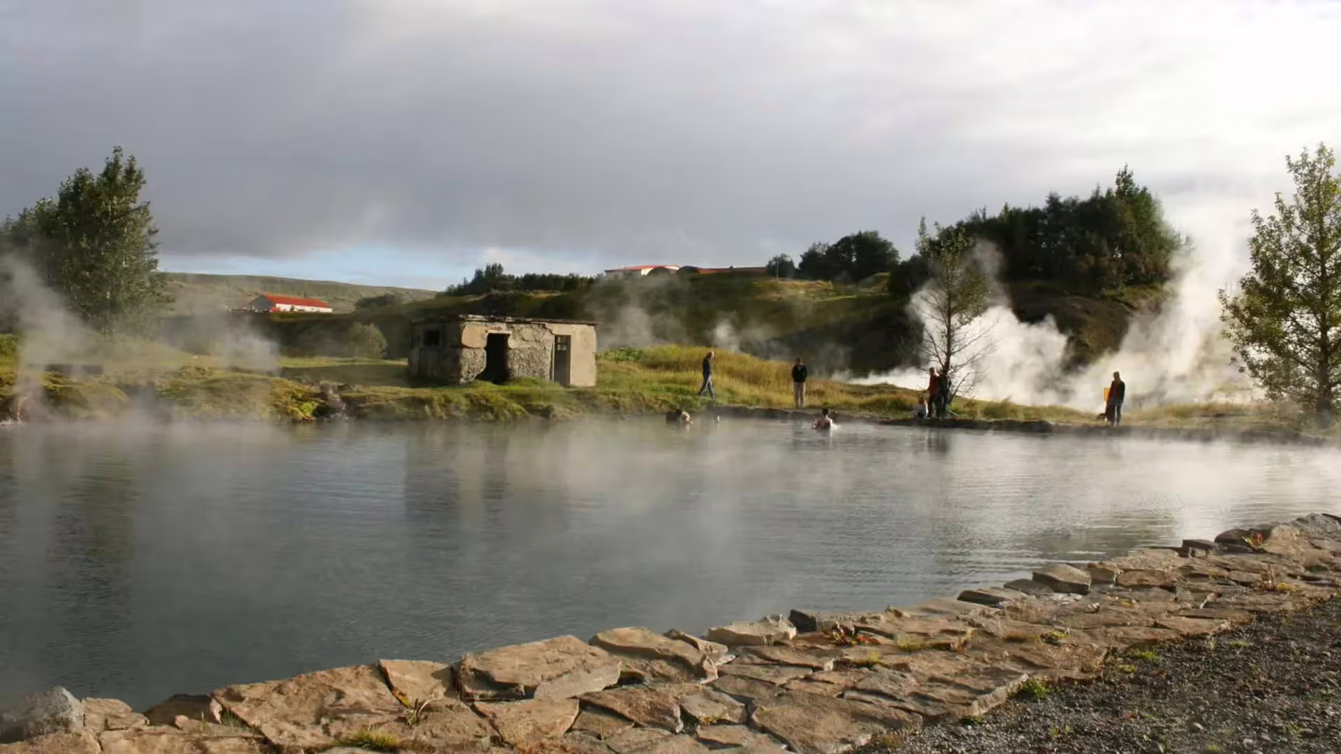 Steaming geothermal Secret Lagoon hot spring pool with visitors and stone building in Iceland's Golden Circle