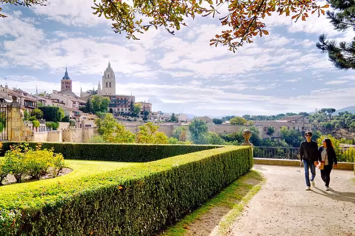 Couple strolls through lush gardens with scenic views of Segovia's historic skyline, a serene stop on the Madrid day tour.