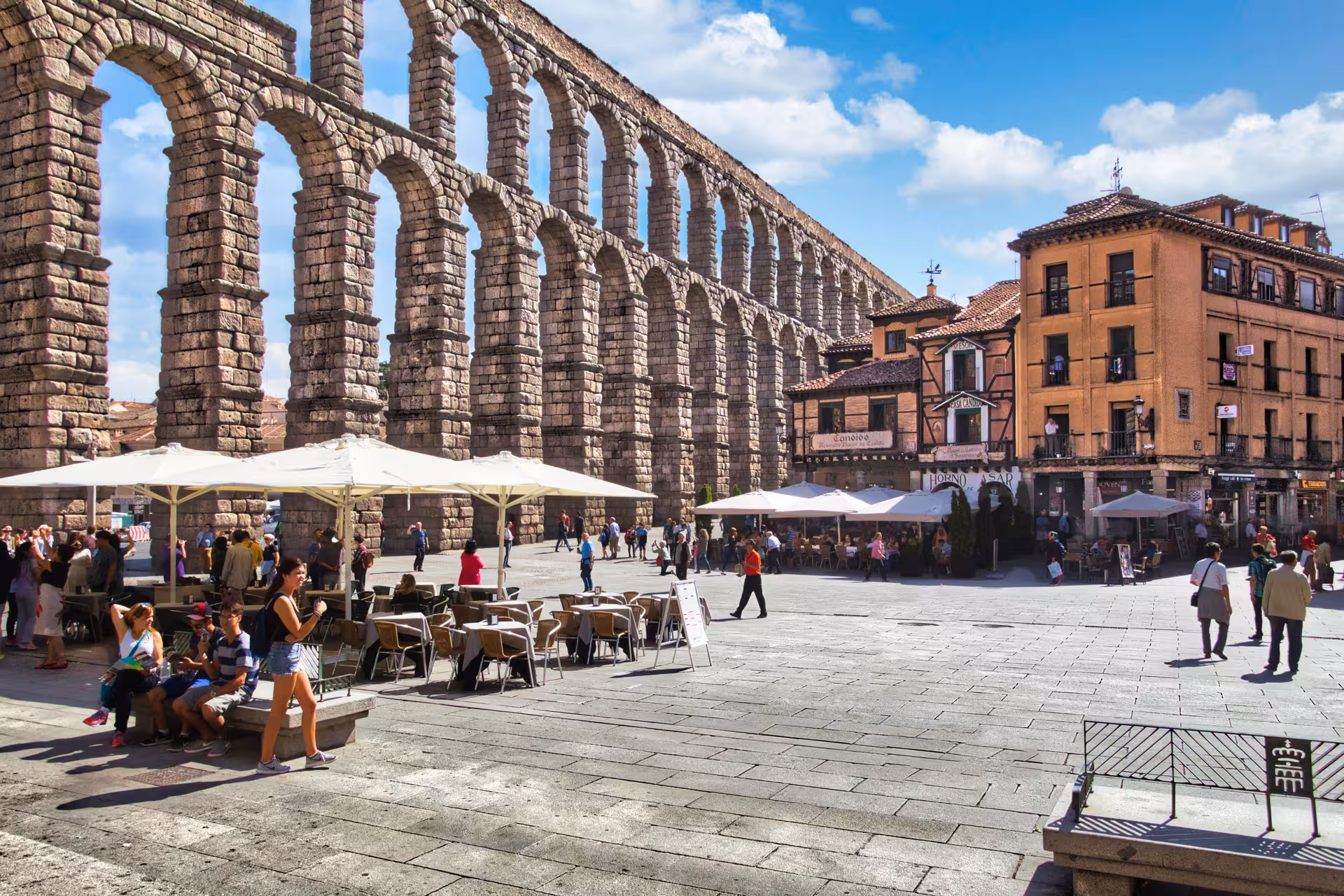 Visitors enjoy outdoor cafes near the iconic Roman Aqueduct in Segovia, a highlight of day trips from Madrid.