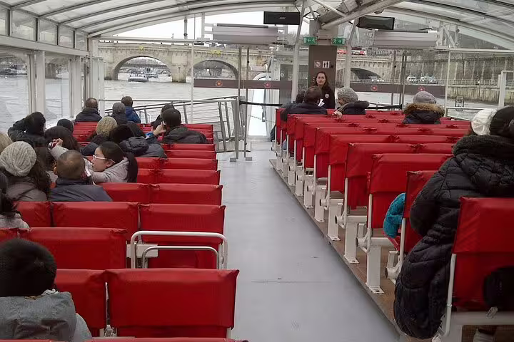 Passengers seated on red chairs inside a Seine River cruise boat with a guide standing at the front