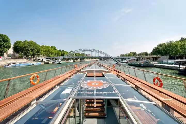 Top deck of a Seine River cruise boat with glass roof, wooden railings, and arched bridge in Paris