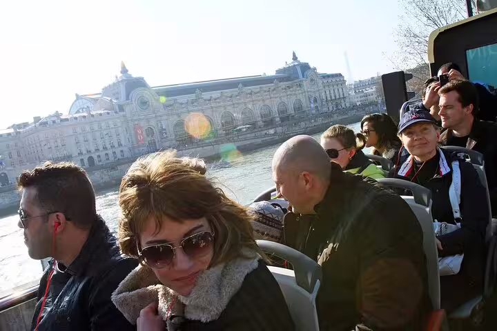 Tourists on open-top hop-on hop-off bus along Seine River with Musée d'Orsay in background, Paris