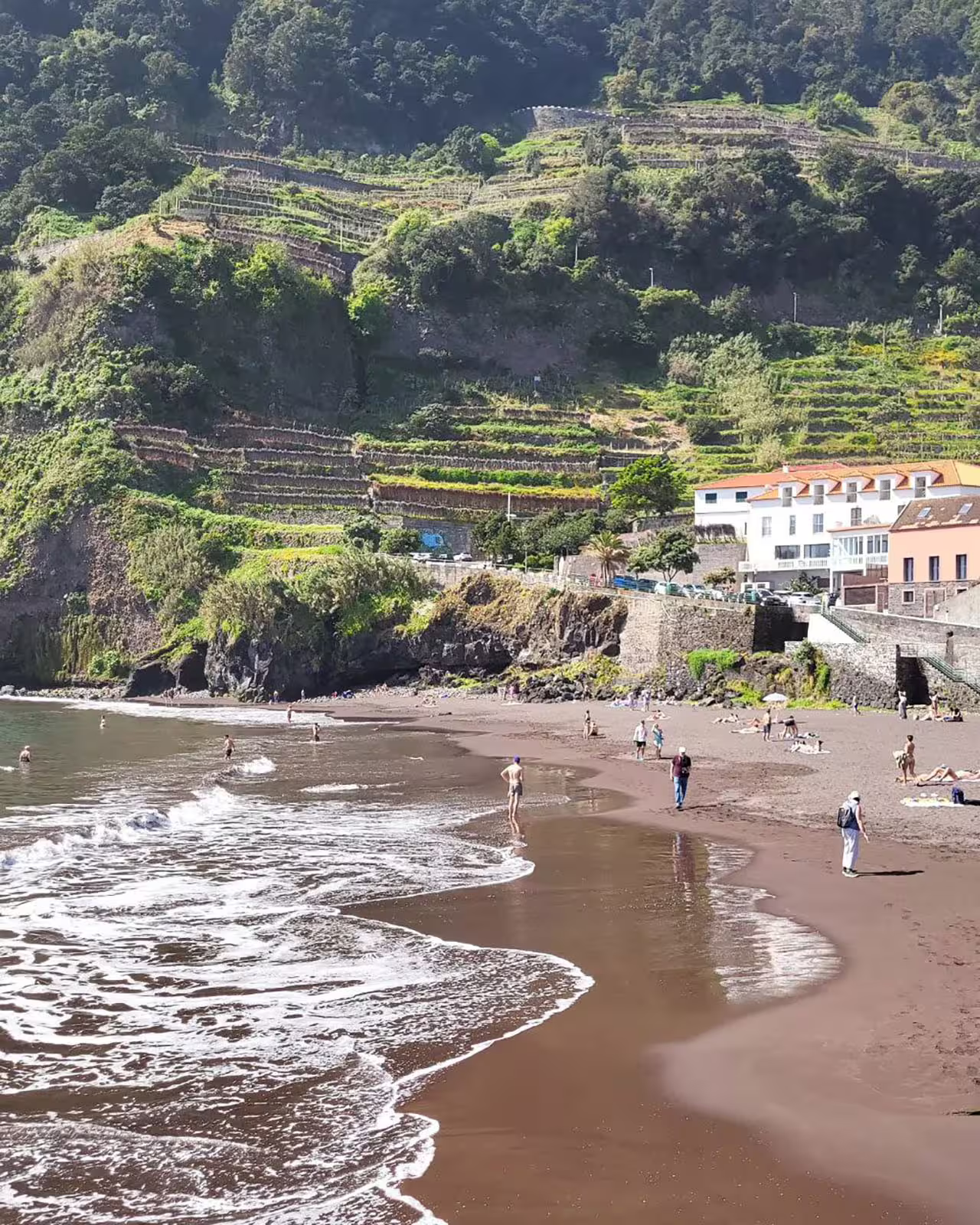 Scenic view of Seixal black sand beach with lush terraced hillsides and visitors enjoying the ocean on Cabo Girão tour.
