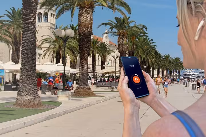 Tourist holding smartphone with Riva audio guide app on Trogir palm-lined waterfront promenade