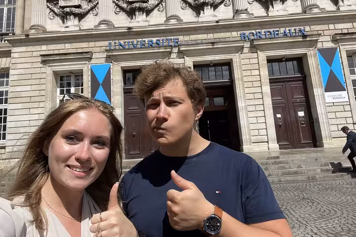 Two tourists giving thumbs up in front of Université Bordeaux historic building on a sunny day