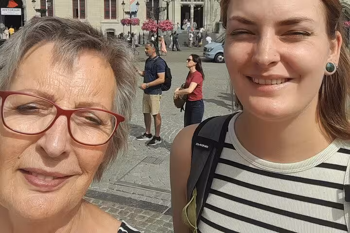 Two women taking a selfie on a cobblestone square in Bruges with historic buildings behind them