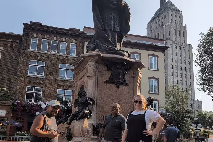 Three tourists posing by a historic bronze statue monument in Old Quebec City with brick buildings behind