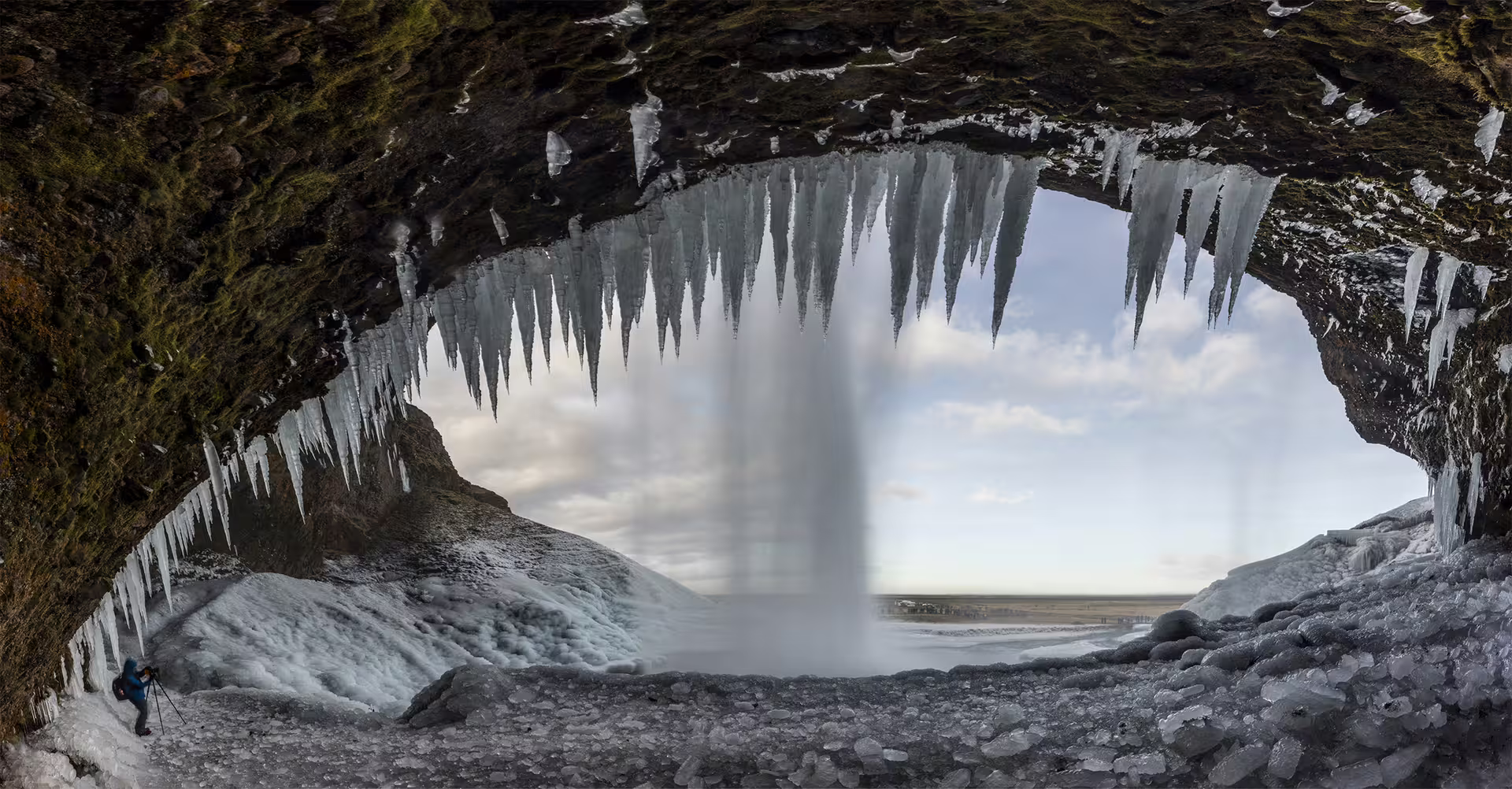 Photographer behind Seljalandsfoss waterfall framed by massive icicles in an ice cave on Iceland's South Coast