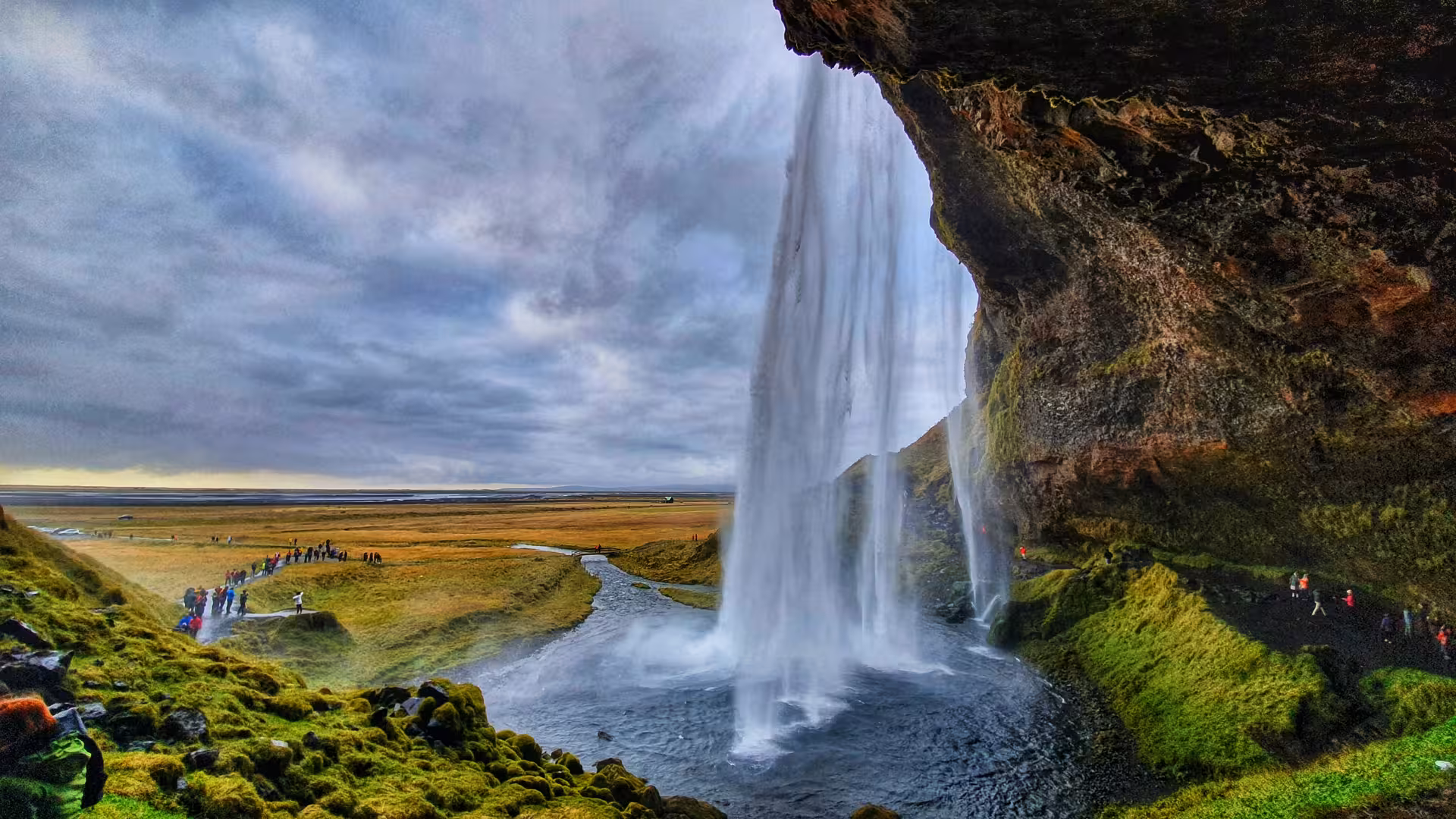 View from behind Seljalandsfoss waterfall in Iceland with visitors on path and golden plains beyond