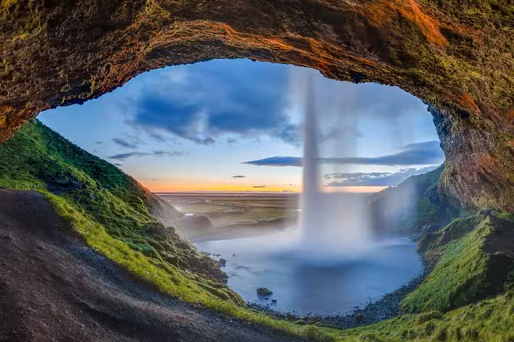 View from behind Seljalandsfoss waterfall in Iceland with lush green cliffs and sunset over the coastal plains