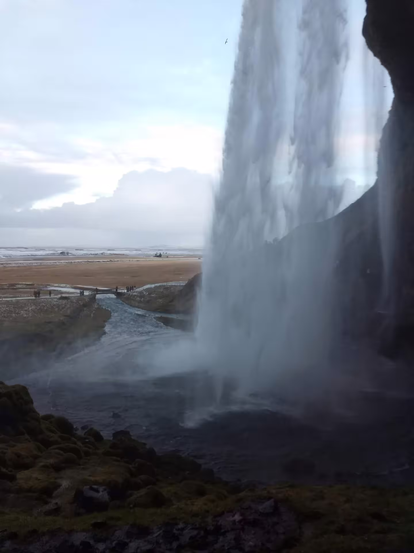 View from behind Seljalandsfoss waterfall in Iceland with misty cascade and flat plains beyond