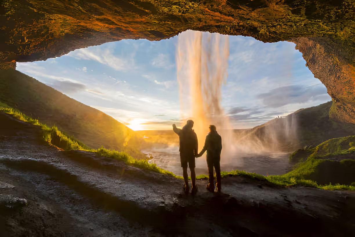 Couple holding hands behind Seljalandsfoss waterfall at golden sunset, viewed from cave, South Coast Iceland