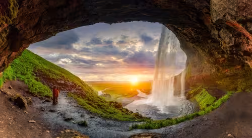View from behind Seljalandsfoss waterfall cave in Iceland at golden sunset with visitor in red jacket