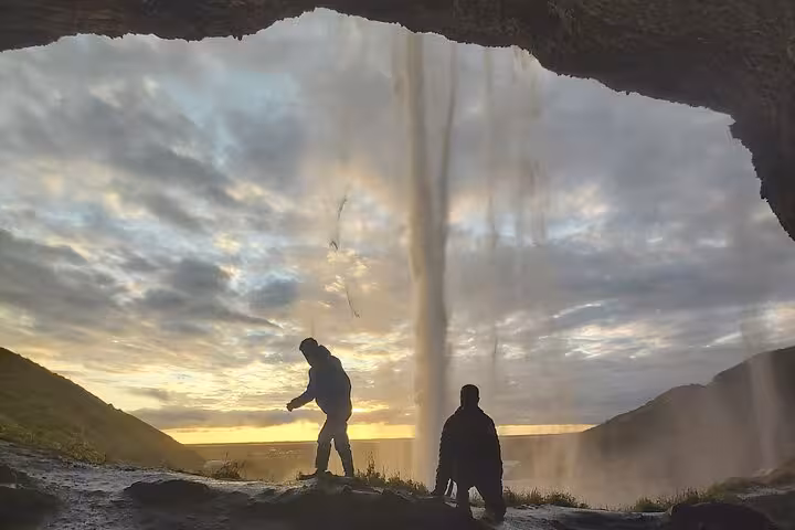 Two silhouetted visitors behind Seljalandsfoss waterfall at golden sunset on Iceland South Coast