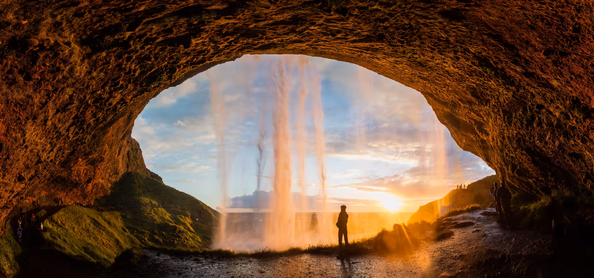 Silhouette of person standing behind Seljalandsfoss waterfall at golden sunset, viewed from cave, Iceland South Coast