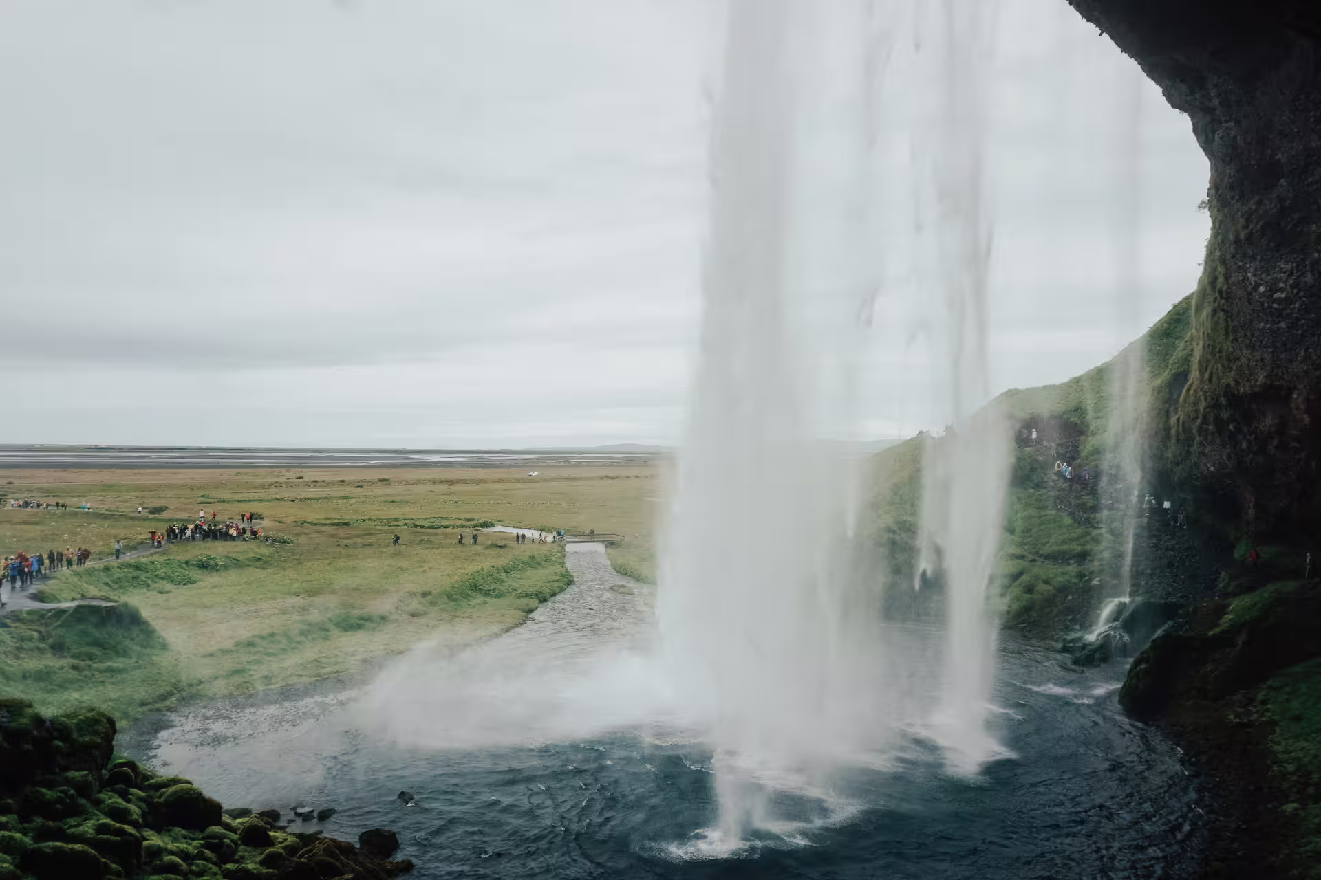 View from behind Seljalandsfoss waterfall in Iceland with vast green plains and tourists visible below