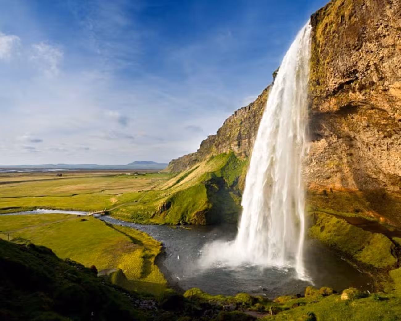 Seljalandsfoss waterfall cascading onto Iceland's green south coast plains under blue sky