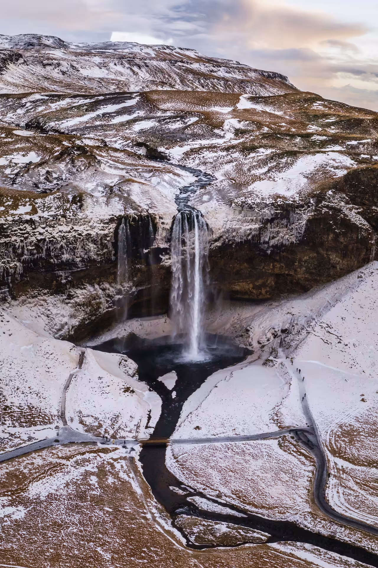 Aerial view of Seljalandsfoss waterfall surrounded by snow-covered cliffs and winding roads in Iceland's South Coast