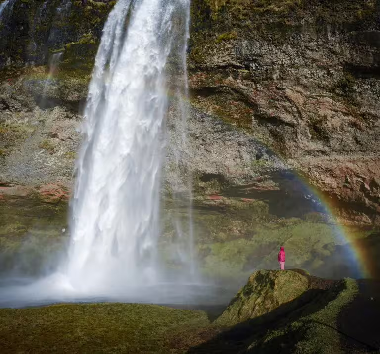 A visitor in pink stands beside a stunning Seljalandsfoss waterfall rainbow on Iceland's South Coast captured private tour