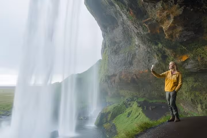 Traveler in yellow jacket taking selfie behind Seljalandsfoss waterfall on Iceland's South Coast