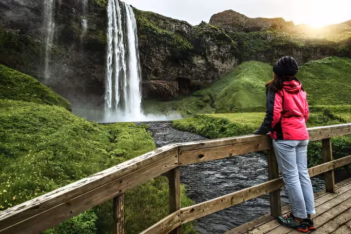 Hiker in pink jacket admiring Seljalandsfoss waterfall from wooden bridge on Iceland South Coast tour
