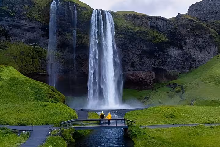 Two visitors in yellow raincoats on a bridge viewing Seljalandsfoss waterfall on Iceland's South Coast