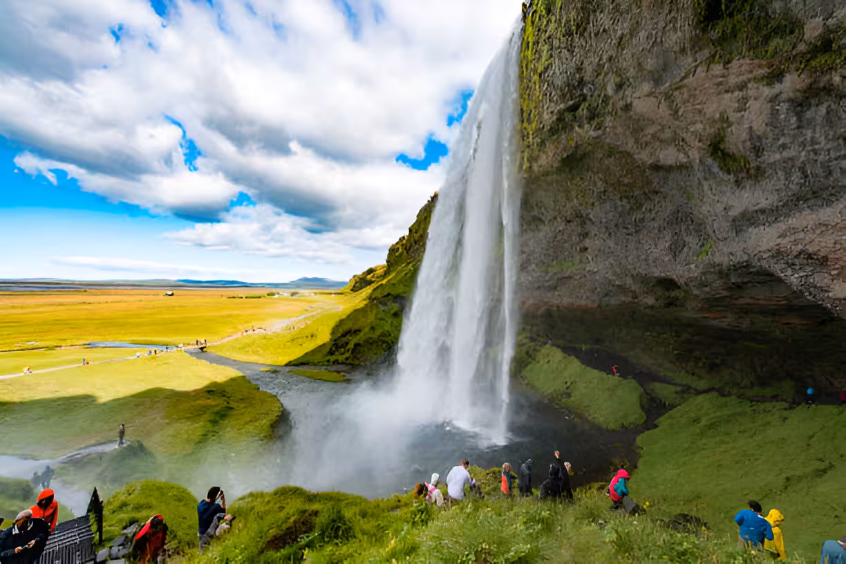 Tourists walking alongside Seljalandsfoss waterfall on Iceland's South Coast with vast green plains beyond