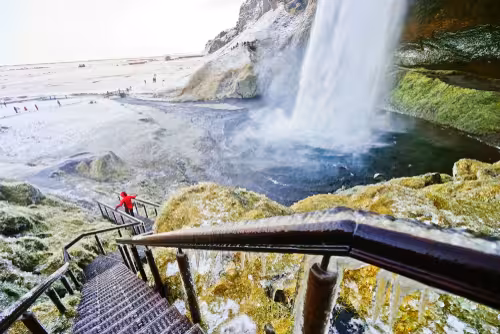 Visitor in red jacket on icy staircase beside Seljalandsfoss waterfall on Iceland South Coast tour