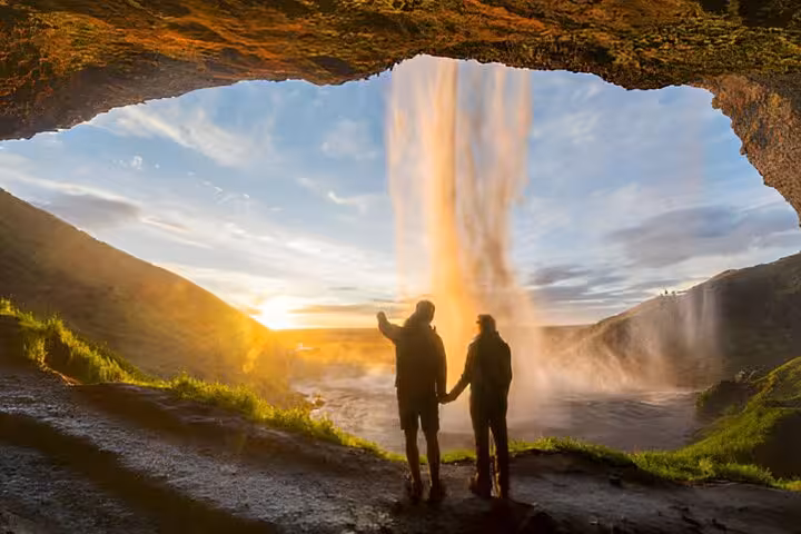 Couple holding hands behind Seljalandsfoss waterfall at golden sunset on Iceland South Coast tour
