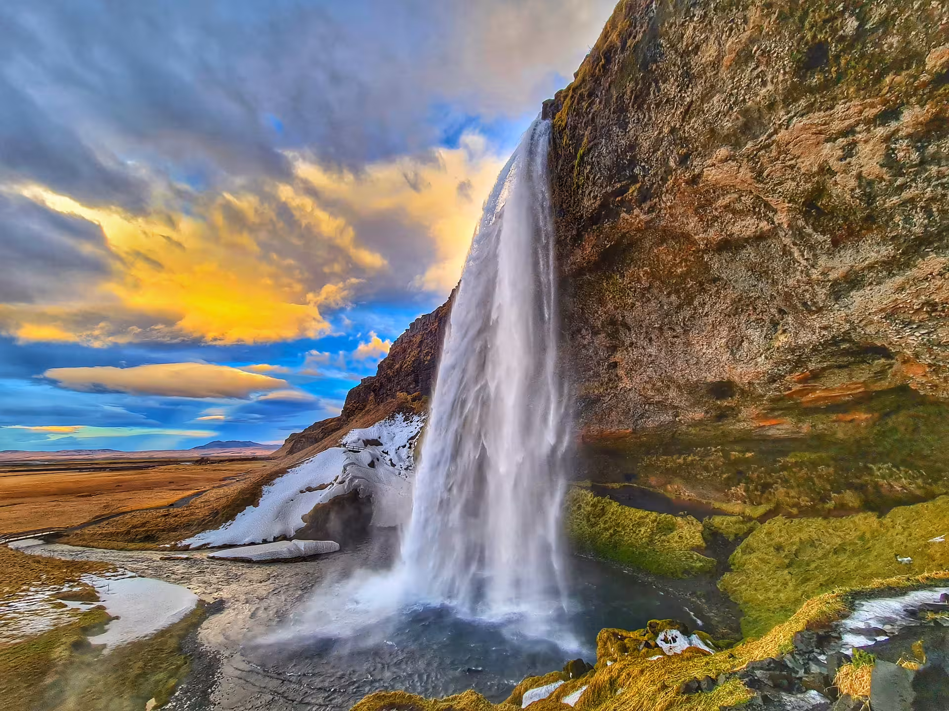 Seljalandsfoss waterfall viewed from behind at sunset with snow and golden clouds, South Coast Iceland