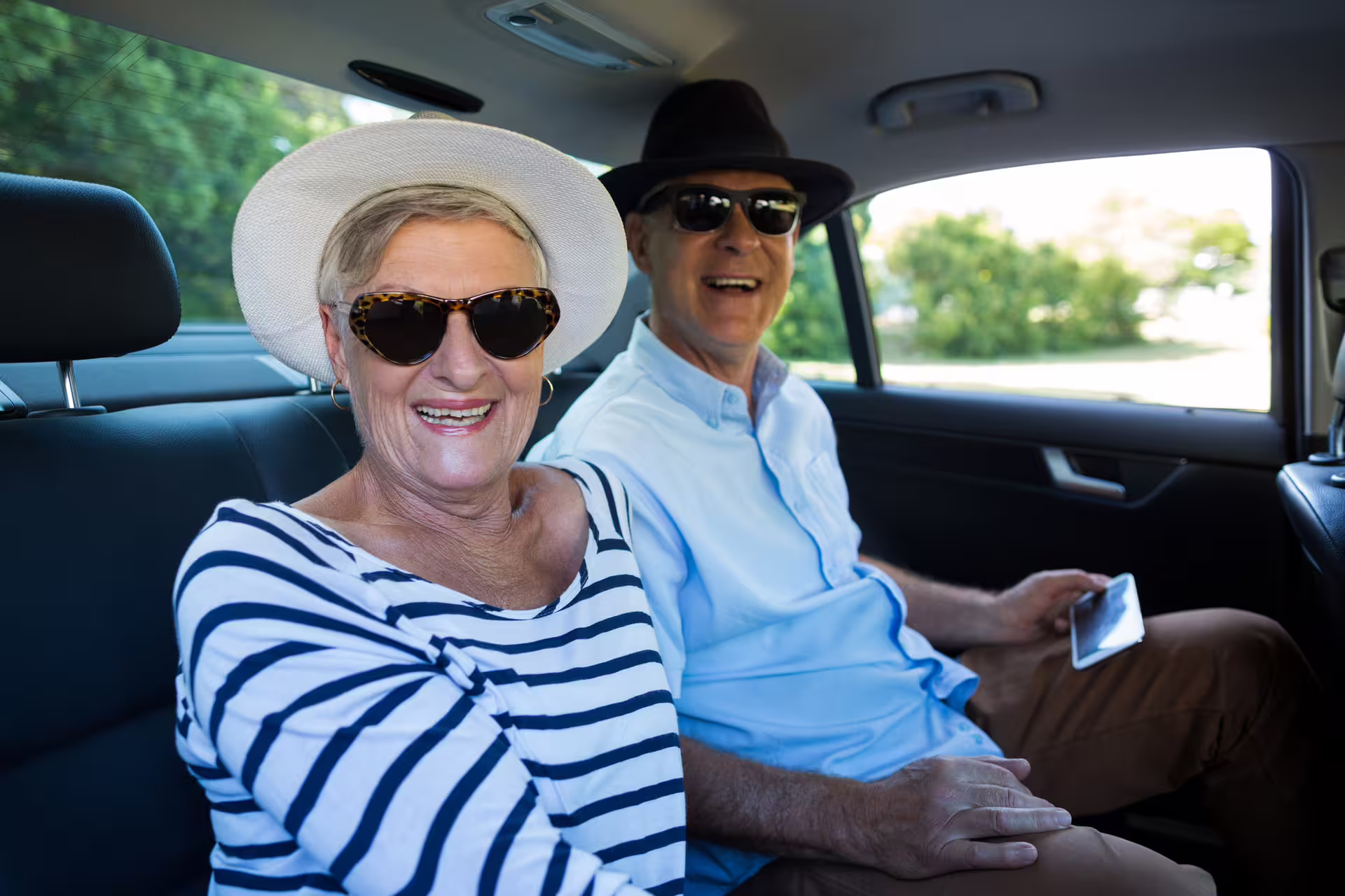 Happy senior couple smiling in back seat of transfer car wearing sunglasses and hats