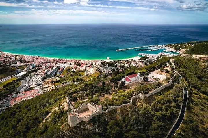 Aerial view of Sesimbra coastline with lush greenery, historic castle, and vibrant blue waters, perfect for a scenic day trip.